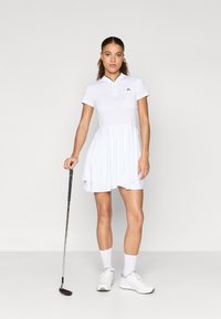 Biała sukienka golfowa z zamek błyskawicznym przy kołnierzyku, krótkimi rękawami i warstwową spódnicą; noszona z białymi trampkami i trzymana w dłoni kij golfowy.