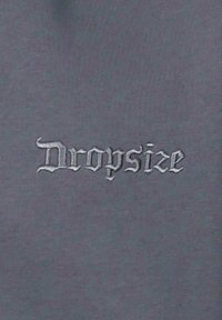 Grauer Stoff mit gesticktem Text "Dropsize" in einer stilisierten Schriftart. Die Textur wirkt glatt und gleichmäßig über die Oberfläche.