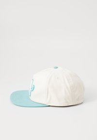 Hvit tekstilhette med lys blå brem. Har et brodert design i blått foran og en matchende knapp på toppen.