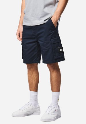 Mann trägt marineblaue Cargo-Shorts mit großen Seitentaschen, weiße Sportsocken und weiße Turnschuhe, steht vor einem schlichten Hintergrund.