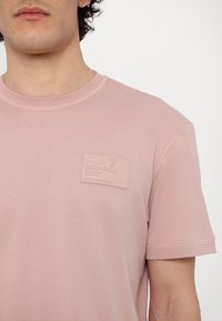 Lys rosa bomull t-skjorte med rund hals, kortermet og en teksturert logo-lapp på brystet. Glatt stoff med en subtil glans.