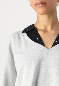 Grå genser med v-utringning, med en svart skjorte under med hvite prikker. Myk tekstur, uformell design.