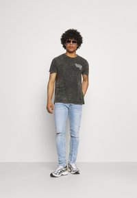 T-shirt cinza desgastada com mangas curtas, apresentando uma gráfica branca no peito. Jeans skinny azul claro com rasgos. Ténis desportivos prateados.