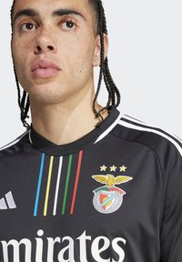 Maglia sportiva nera con righe verticali verdi, gialle e blu. Presenta un logo del club e testo bianco, con un design a colletto a V.