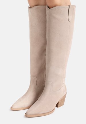 Beige suède knie hoge laarzen met spits toelopende neuzen en gestapelde houten hakken gedragen aan benen tegen een effen achtergrond.