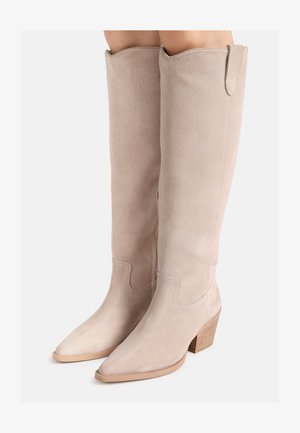 Bottes hautes jusqu'aux genoux en daim beige avec bouts pointus et talons en bois empilés, portées sur des jambes devant un fond uni.