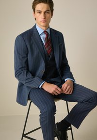 Costume bleu marine à rayures avec deux boutons, gilet assorti, chemise bleu clair, cravate rouge à rayures, assis sur une chaise en métal noir.