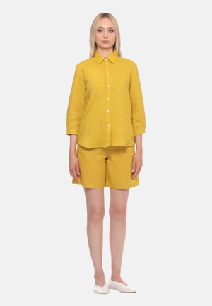 Donna in piedi rivolta in avanti, indossa una camicia giallo senape con bottoni, shorts abbinati e scarpe basse bianche su uno sfondo semplice.