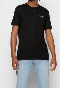 Černé pánské tričko s krátkým rukávem typu Henley s ribovanou texturou, vybavené třemi knoflíky a bílým logem "Calvin Klein Jeans" na hrudi.