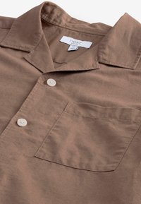 Brun kortærmet skjorte lavet af blødt stof, med en notch krave, to hvide knapper og en brystlomme. Farven er ensartet med minimal tekstur.