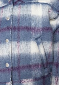 Rutigt flanellskjorta i nyanser av blått, vitt och lila. Har metalliska tryckknappar och en sidficka på framsidan. Mjuk textur.