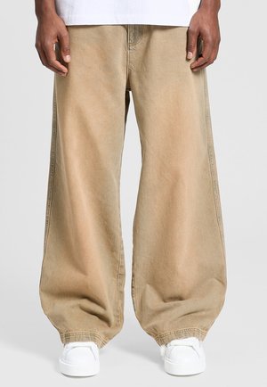 Person iført løstsiddende beige bukser og hvide sneakers, stående mod en ensfarvet lysegrå baggrund.