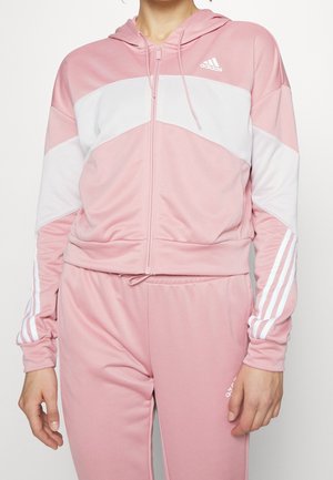 Persoon die een roze Adidas trainingspak draagt met witte chevron en strepen, een hoodie met ritssluiting en bijpassende broek, getoond van nek tot halverwege de dij.