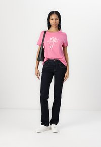 Rosa bomulls-t-skjorte med hvitt blomstermotiv, kombinert med mørkeblå jeans og hvite sneakers. Modell som holder en liten svart veske.