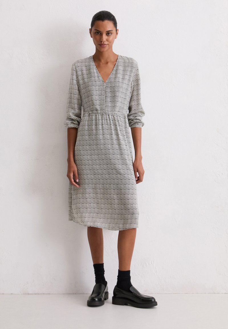 Schwarz-weiß gemustertes Kleid mit V-Ausschnitt, langen Ärmeln und elastischem Bund, kombiniert mit schwarzen Socken und Schuhen.