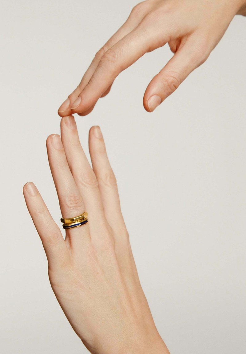 Tres anillos metálicos apilados en un dedo; uno dorado, uno plateado y uno negro. Manos que se extienden una hacia la otra con tonos de piel neutrales.