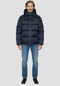 Veste matelassée bleu foncé avec capuche, comportant des sections matelassées horizontales, une fermeture éclair complète et un logo sur le côté gauche de la poitrine. Portée avec un jean bleu.