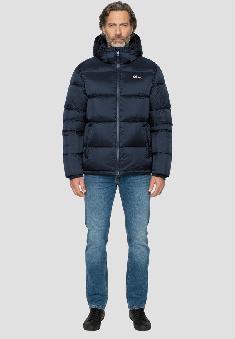 Veste matelassée bleu foncé avec capuche, comportant des sections matelassées horizontales, une fermeture éclair complète et un logo sur le côté gauche de la poitrine. Portée avec un jean bleu.