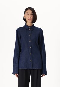 Tumma sininen farkkupaita, jossa on kauluksesta, pitkät hihat ja napitus. Omavoimainen leikkaus ja leveät hihansuut.