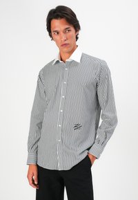 Striped black and white button-up shirt with a white collar, featuring long sleeves and a small graphic detail on the lower front.