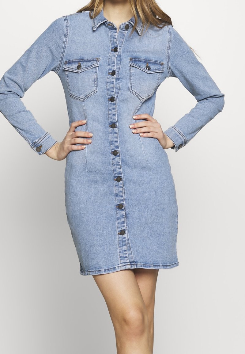 Vestido de denim en azul claro, con mangas largas, frontal abotonado, dos bolsillos en el pecho y una silueta ajustada con una longitud hasta medio muslo.