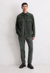 Zielona kurtka sztruksowa z dwoma kieszeniami na piersi, w komplecie z dopasowanymi spodniami i czarnymi sznurowanymi butami. Model stoi na jednolitym tle.