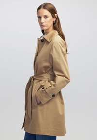 Beige trenčkot s opaskou v pase, knoflíkovými manžetami a límcem. Vyroben z hladké látky, má dvě boční kapsy a volnější střih.