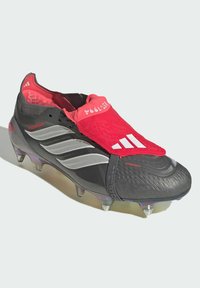 Taco de fútbol en negro y rojo con parte superior sintética texturizada, cuello elástico tipo calcetín, detalles en gris y tachones transparentes para tracción.