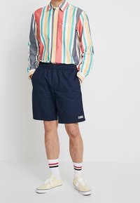 Camicia a righe con linee verticali multicolori, pantaloni corti blu navy, calzini bianchi con righe rosse e nere, e scarpe da ginnastica gialle.
