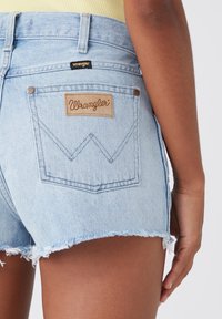 Světle modré džínové kraťasy s třepeným lemem, s hnědou nášivkou „Wrangler“ a charakteristickým vzorem na zadní kapse.