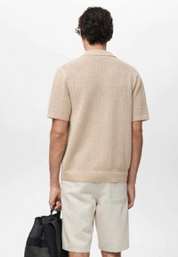 Beige knitted short-sleeve polo shirt with a textured pattern, paired with light-colored shorts. The individual holds a black bag.