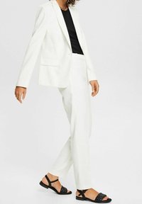 Biały, dopasowany żakiet i pasujące spodnie z gładkiej tkaniny, zestawione z czarnymi, płaskimi sandałami z plecionymi paskami.