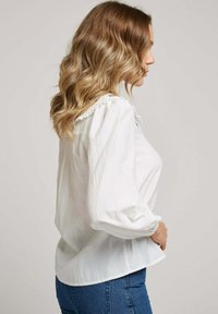 Blouse blanche à manches longues avec encolure froncée et poignets resserrés, présentant une coupe décontractée et une texture douce, associée à un jean bleu ajusté.