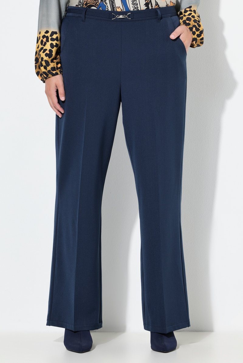 Vrouw draagt marineblauwe, op maat gemaakte broek met metalen gespsluiting, luipaardprint en grafische blouse met lange mouwen, en marineblauwe puntige schoenen.