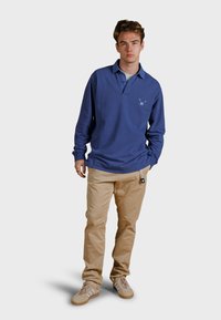 Blå långärmad polo med krage, logotyp på bröstet, tillsammans med beigefärgade byxor och bruna sneakers, som visar en avslappnad outfit.