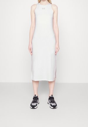 Femme debout portant une robe midi blanche sans manches avec le logo EA7 et des baskets chunky noires et blanches sur un fond blanc uni.