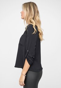 Schwarzes Langarmshirt mit einer Fronttasche, lockerem Schnitt, aufgekrempelten Ärmelabschlüssen, weicher Textur und geradem Saum. Kombiniert mit eng anliegenden Leggings.