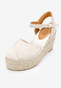 Sandalias de cuña beige con parte superior de ante y plataforma tejida de yute. Correa ajustable en el tobillo con hebilla metálica y tacón texturizado.