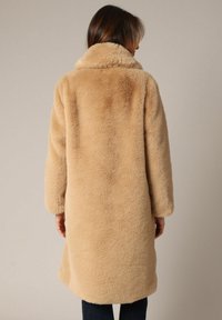 Abrigo de piel sintética beige con cuello alto, mangas largas y un diseño de longitud media, que presenta una superficie suave y texturizada.