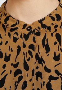 Brauner Stoff mit schwarzem Leopardenmuster, mit Stehkragen und sichtbaren Knopfdetails, die eine glatte, strukturierte Oberfläche hervorheben.
