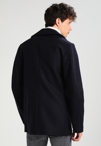 Cappotto di lana blu navy con design strutturato, ampi rever e silhouette pulita. Presenta una texture liscia e cucitura posteriore dritta.