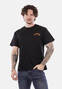 T-shirt in cotone nero con maniche corte, collo rotondo e testo arancione "PROPAGANDA" sul petto. Adatto per un abbigliamento casual.