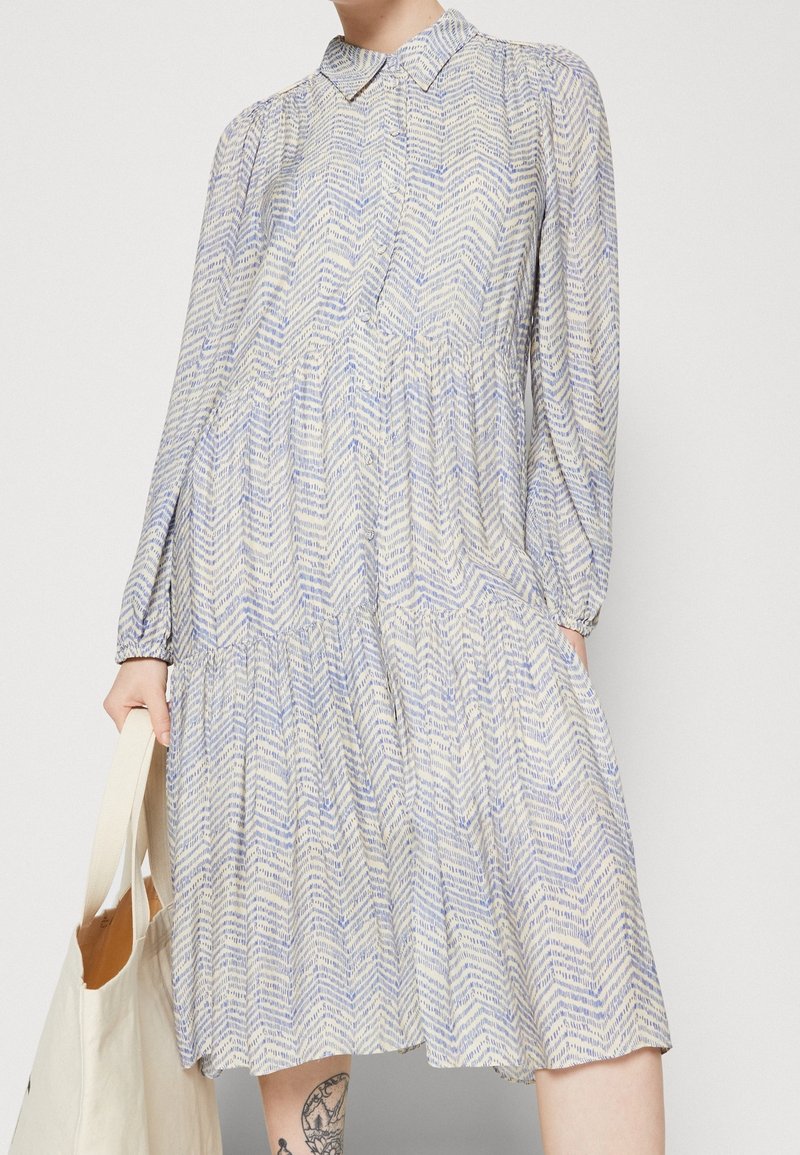 Robe à manches longues, longueur genou, motif chevron bleu et beige, boutonnée sur le devant, col de chemise, portée avec un sac fourre-tout blanc et une jambe tatouée visible.