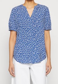 Blouse bleue à manches courtes avec un motif floral texturé en blanc. Présente un décolleté en V et un ourlet légèrement incurvé pour une coupe décontractée.