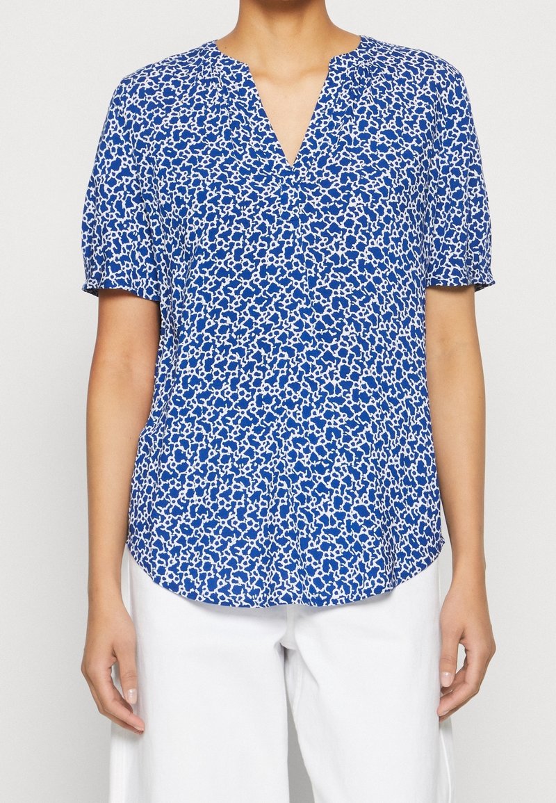 Blouse bleue à manches courtes avec un motif floral texturé en blanc. Présente un décolleté en V et un ourlet légèrement incurvé pour une coupe décontractée.