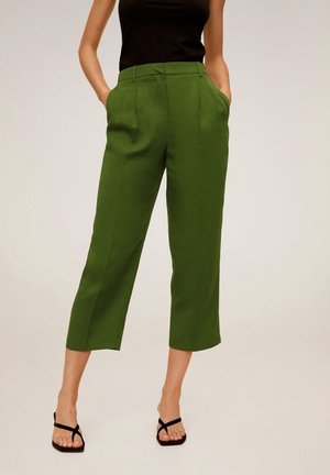 Vrouw draagt olijfgroene, hooggetailleerde, cropped broek met voorplooien, gecombineerd met zwarte sandalen met bandjes en een mouwloos zwart topje.