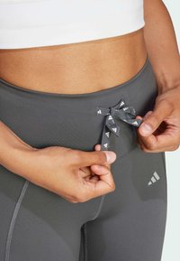 Hands adjusting the drawstring bow on grey athletic leggings worn with a white cropped top, showing midriff and waistband details.