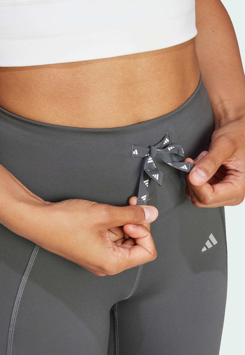 Hände justieren den Kordelzug an grauen Sportleggings, die mit einem weißen Cropped-Top getragen werden und den Bauch sowie die Details des Bunds zeigen.