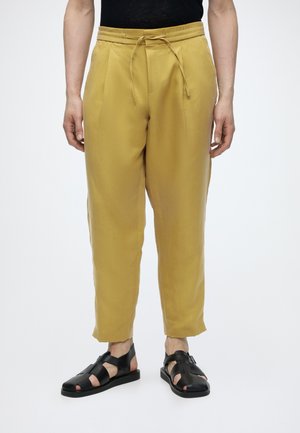 Personne portant un pantalon à cordon de serrage jaune moutarde et des sandales en cuir noir, debout sur un fond blanc.