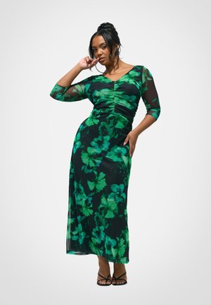 Femme portant une longue robe noire à motifs floraux verts, manches trois-quarts, devant froncé, posant avec une main près du visage et l'autre sur la hanche.